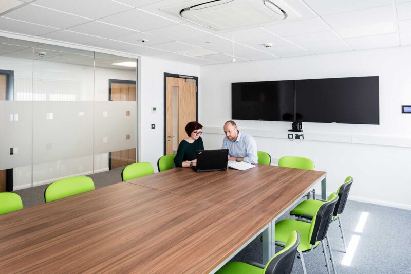 Meeting room inside a modular office building with two people reviewing work at a wooden table with green chairs and a screen.